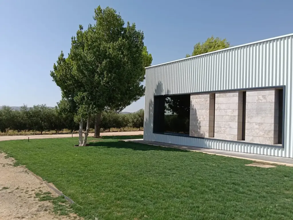 Fachada exterior de la sala de catas de Pagos de Aherca rodeada de césped, pinos y olivar en Valdepeñas